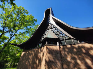 An exterior view of the Agudas Achim sanctuary shows a sharply peaked roof with flared eaves, and the brick walls and patterned glass of the building.